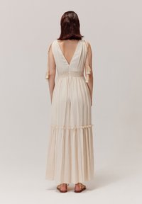 Maxi sukienka w jasnobeżowym kolorze, z głębokim dekoltem na plecach, marszczonym pasem i warstwową spódnicą z falbankami. Detale z frędzlami na ramiączkach.