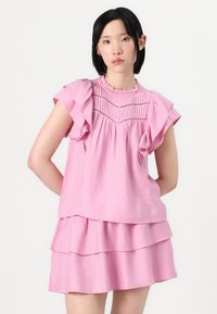 Robe rose courte avec des manches volantes, un buste texturé et une jupe superposée. Elle présente un décolleté froncé et des détails en dentelle.