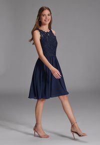 Marineblaues Kleid mit einem Spitzenoberteil, das mit aufwendiger Stickerei verziert ist, einer taillierten Taille und einem fließenden Chiffonrock. Getragen mit nude-farbenen Riemchenheels.