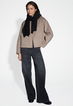 Beige cropped jack met twee voorszakken, gecombineerd met donkere spijkerbroek en een zwarte, gestructureerde sjaal, staand tegen een lichte achtergrond.