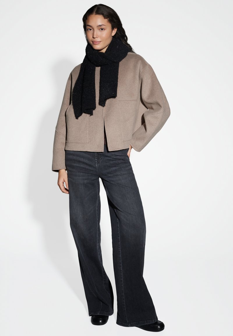 Giacca beige corta con due tasche frontali, abbinata a jeans scuri e una sciarpa nera lavorata, in piedi davanti a uno sfondo chiaro.