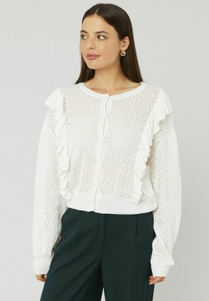 Gilet en tricot blanc avec un motif en losanges texturé, présentant des accents volants le long de l'avant. Fermeture à boutons et manches longues.