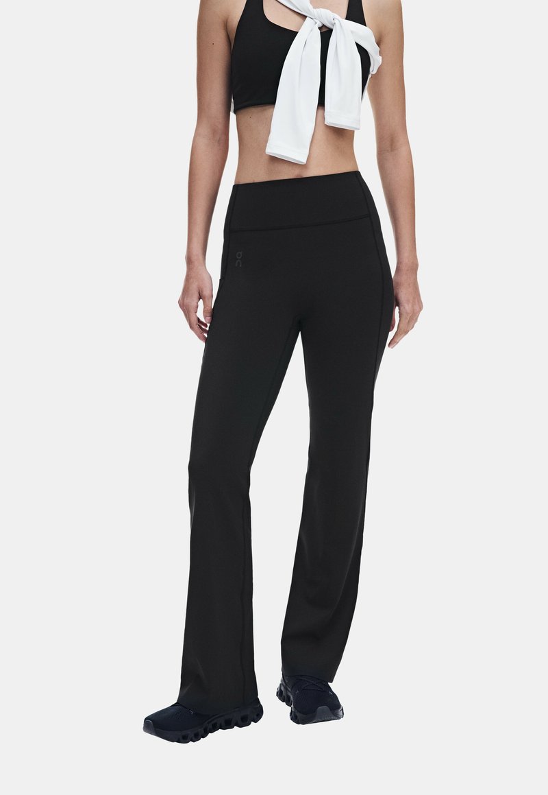 Femme portant des leggings évasés noirs, un soutien-gorge de sport noir, des baskets noires et un haut à manches longues blanc noué autour du cou.