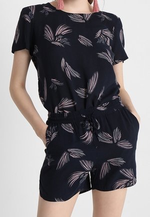 Vrouw draagt een zwarte korte mouwen top en short set met roze veerachtige patronen en elastische taille met trekkoord.