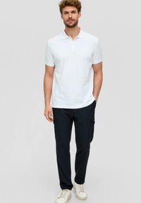 Witte polo met kraag, gecombineerd met zwarte cargobroek en lichtgekleurde sneakers. Model staat tegen een effen grijze achtergrond.