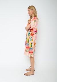 Bunt bedrucktes Blumenkleid mit kurzen Ärmeln, aus leichtem Stoff, mit leuchtenden Mustern und geometrischen Akzenten. Kombiniert mit Riemchensandalen.