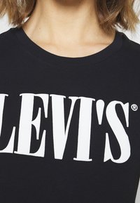 Gros plan du torse portant un t-shirt noir Levi's avec un grand logo blanc Levi's sur la poitrine, l'encolure et une partie des cheveux courts visibles.