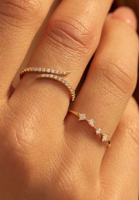 Deux bagues en or sur une main. L'une a un bandeau ondulé orné de petites pierres transparentes, tandis que l'autre présente trois pierres carrées transparentes en rang.