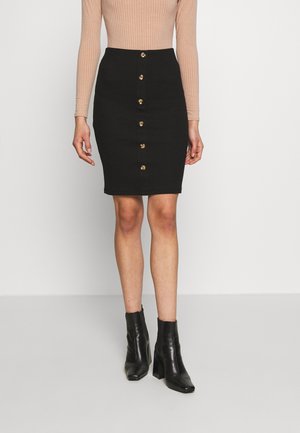 Vrouw draagt een zwarte rok met knopen aan de voorkant, een beige geribd shirt met lange mouwen en zwarte enkellaarzen, staand tegen een effen witte achtergrond.