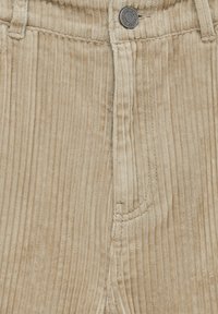 Beežid kõrvitsad, millel on vertikaalne ribastus, eesmine nööpkinnitus ja küljetasku, millel on kontrastne õmbluse üksikasjad.