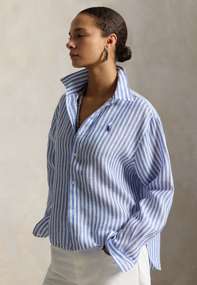 Kvinne med hår i knute iført blå og hvit stripete skjorte med knapper og hvite bukser, stående i et nøytralt farget miljø, med hendene i lommene.