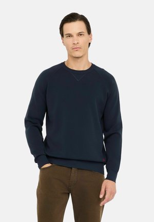 Felpa color navy con scollatura a girocollo, polsini e orlo a coste, caratterizzata da un sottile dettaglio a triangolo cucito sul collo. Abbinata a pantaloni marroni.