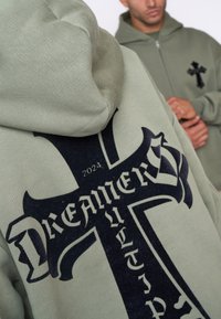 Felpa verde in misto cotone, con una grande stampa nera a croce e il testo "DREAMERS". Tessuto liscio, vestibilità comoda e cappuccio.