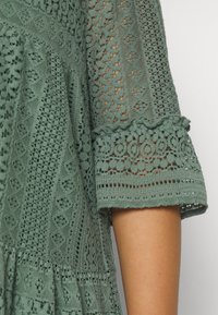 Grünes gehäkeltes Kleid mit komplizierten Mustern, strukturiertem Stoff, weit ausgestellten Ärmeln mit gewelltem Saum und mehrlagigem Design.