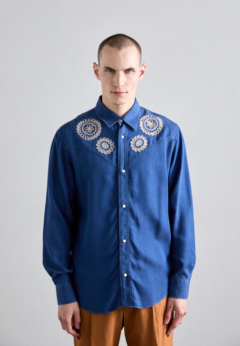 Dunkelblaue Jeanshemd mit langen Ärmeln, das bestickte, kreisförmige Designs auf den Schultern sowie eine Knopfleiste hat.
