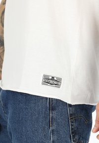 T-shirt bianco in cotone con orlo arrotondato e un'etichetta nera cucita che recita "DENIM FACTORY." Abbinato a jeans in denim blu.