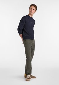 Sudadera marina con cuello redondo, combinada con pantalones verde oliva y zapatillas marrones con acentos amarillos y cordones texturizados.