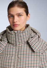 Pufasta jakna s visokim ovratnikom, houndstooth uzorak u bež, smeđoj i tamnoplavoj boji, s teksturiranim završetkom i vidljivim šavovima.