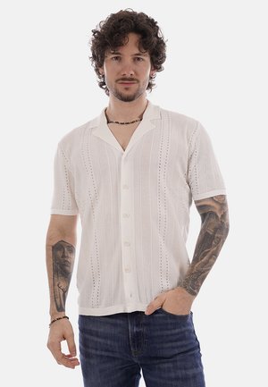 Homme aux cheveux bouclés portant une chemise blanche à manches courtes avec motifs et un jean foncé, montrant des tatouages sur les deux bras.