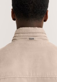 Beige suede jacket with a folded collar, featuring a small metal logo tag on the back, and visible stitching along the seams.