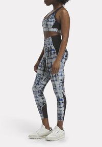 Conjunto de sujetador deportivo y leggings en gris con patrón abstracto en azul y negro, fabricado en tela elástica con detalles en negro, combinado con zapatillas blancas.