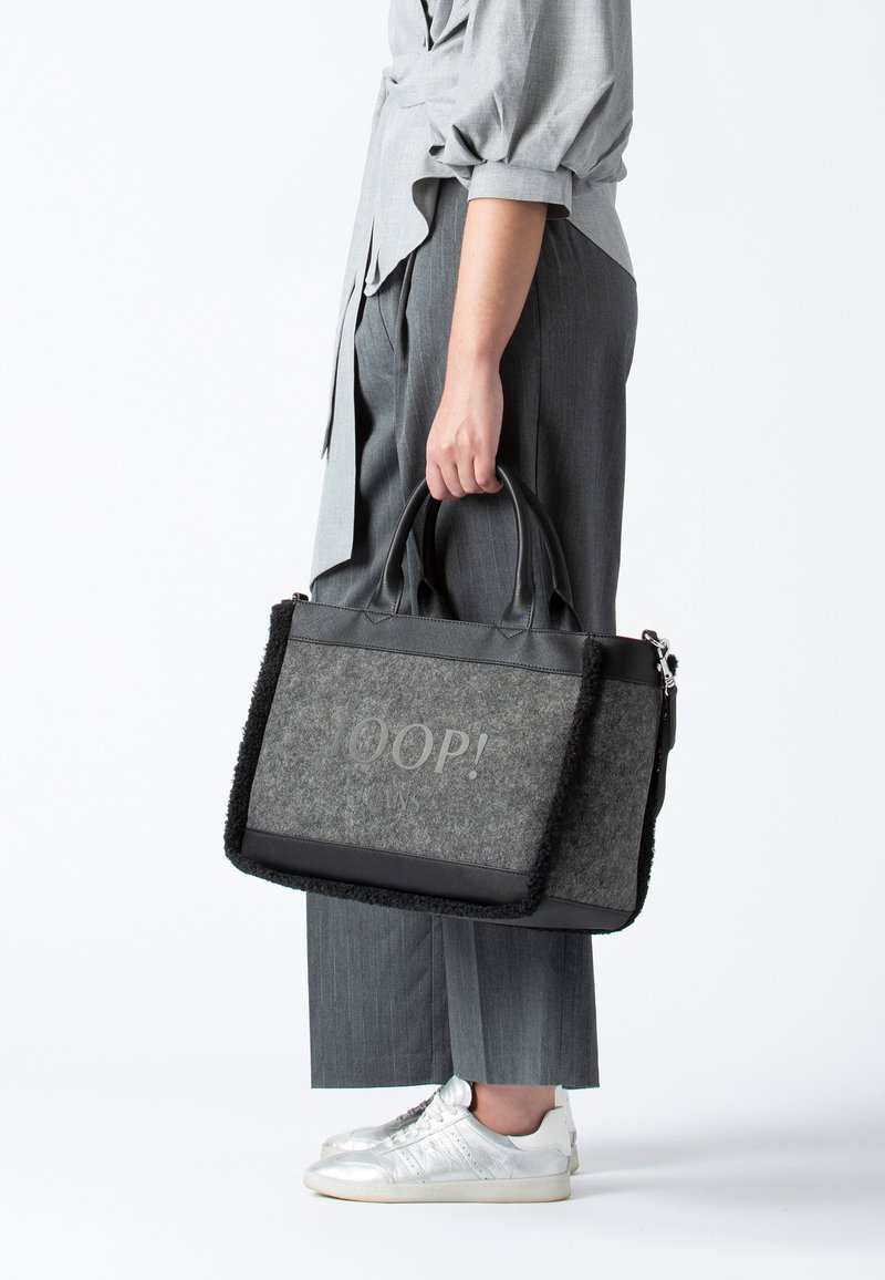 Grå handväska med svarta läderdetaljer, med texten "OOP!" framtill och en luddig kant. Kort handtag och justerbar axelrem.