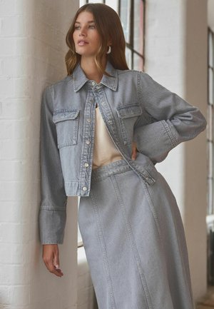 Svetlo sivá džínová bunda s prednými vreckami a zapínaním na gombíky, v kombinácii s pasujúcou sukňou. Hladká textura a jednoduchý dizajn, umiestnený proti múru.