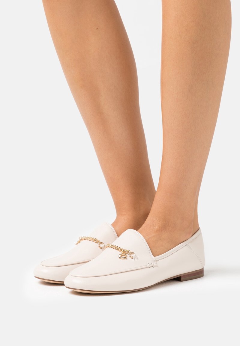 Pieds chaussés de mocassins en cuir blanc avec un détail de chaîne dorée, debout devant un fond clair uni.