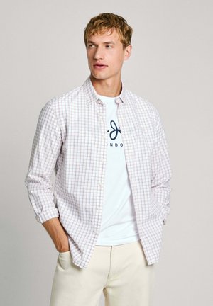 Chemise blanche à carreaux avec un motif de grille bleu clair et orange, avec des poignets boutonnés, portée sur un t-shirt blanc affichant un texte bleu marine, associée à un pantalon beige clair.