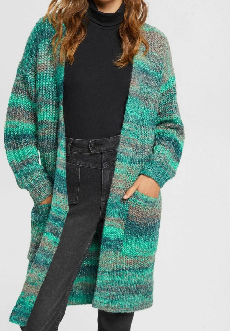Długi kardigan zapinany z przodu w kolorze teal i szarym, o fakturowanym wzorze dzianiny. Posiada dwie kieszenie z przodu oraz ściągacze na mankietach. Noszony na czarną bluzkę.