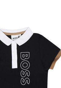 Polo noir avec col et boutons blancs, arborant un logo "BOSS" blanc en gras sur le devant. Accents marron sur les bords des manches. Matière en coton.