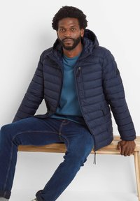 Navy puffer jacket with a hood, featuring horizontal quilted texture, zip pockets, and a logo on the left chest, paired with a blue shirt and jeans.