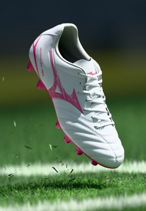 Wit-roze voetbalschoen met het label "Monarcida Neo" die gras opwaait op een voetbalveld nabij een witte lijn.