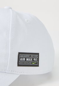 Casquette blanche en tissu avec un design structuré. Présente une étiquette noire affichant "AIR MAX 95" en blanc et des accents jaune fluo.