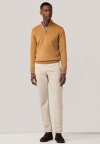Pull en moutarde avec un col zippé, associé à un pantalon beige clair. La tenue comprend une chemise à carreaux bleus et blancs en dessous.