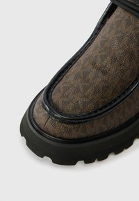 Közelkép egy barna textúrájú loafer cipőről fekete díszítéssel és vastag fekete gumi talppal, fehér háttéren.