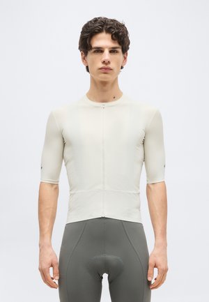 Noor mees, kes kannab keha järgivat beeži rattasärki esilukuga ja hallide polstriga rattapükse, seisab tavalise tausta ees.