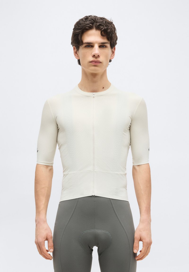 Νέος άνδρας που φοράει εφαρμοστή μπεζ φανέλα ποδηλασίας με φερμουάρ στο μπροστινό μέρος και γκρι επενδυμένα σορτς ποδηλασίας, στέκεται μπροστά σε απλό φόντο.