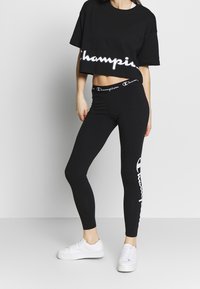 Czarny krótki t-shirt z logo "Champion" nadrukowanym na biało, w połączeniu z czarnymi legginsami z białym logo "Champion" z boku. Białe tenisówki.