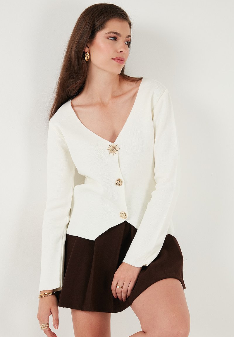 White knit cardigan with V-neck, featuring a gold sun-shaped button at the neckline and three gold buttons down the front, paired with a brown skirt.