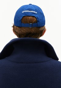 Person mit kurzen braunen Haaren trägt eine blaue Mütze mit dem Text "ARMEDANGELS" und einen hochkragen navyblauen Pullover, von hinten betrachtet.