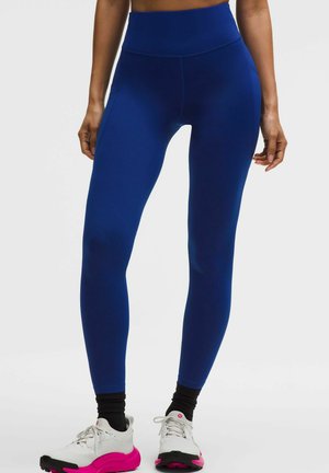 Frau trägt hoch taillierte marineblaue Leggings, schwarze Socken und weiße Laufschuhe mit rosa Sohlen, steht vor einem schlichten Hintergrund.