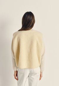 Šviesiai geltonas vilnonis megztinis su tekstūruota paviršiaus danga, laisvo silueto, su minkštais ir ilgais rankovėmis. Derinamas su baltais džinsais, matomas iš nugaros.