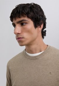 Beige gebreide trui met ronde hals, een gestructureerde afwerking en een klein logodetail op de borst; gedragen over een witte T-shirt kraag.