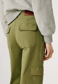 Persoon in olijfgroene cargobroek met klepzakken, een bruine leren merkembleem en een lichtgrijze sweatshirt.