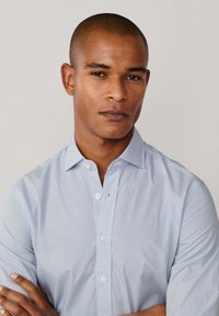 Lichtblauw overhemd met subtiele geometrische patronen, met kraag en lange mouwen. Heeft witte knopen en een gestructureerde stof.