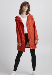 Giacca impermeabile arancione brillante con cappuccio, bottoni a pressione, tasche laterali e una vestibilità leggermente rilassata, abbinata a pantaloni neri e sneakers bianche.