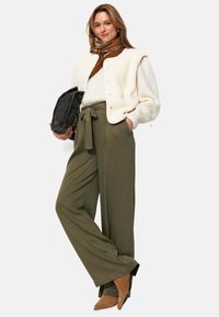 Gilet sans manches en fausse fourrure crème sur un haut marron, pantalon large olive, et bottes à talons marron. Veste en denim noire tenue d'une main.