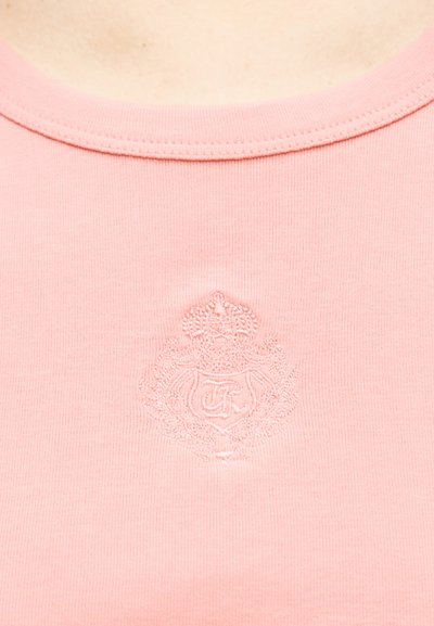 Κοντινό σχέδιο ροζ υφάσματος τοπ με διακριτικό κεντητό έμβλημα που περιλαμβάνει στέμμα, στεφάνι δάφνης και ασπίδα με διαπλεκόμενα αρχικά.