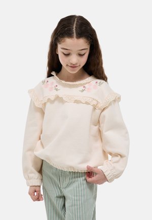 Jeune fille aux longs cheveux portant un pull crème avec broderie florale et dentelle à volants, associé à un pantalon rayé vert et blanc.